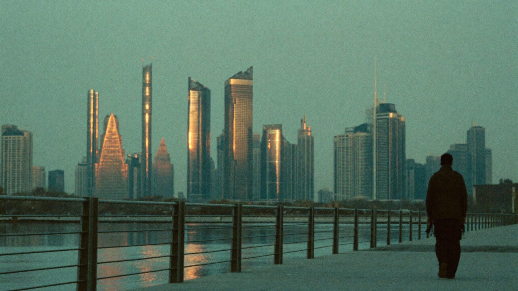 A man walking a dog along Dubai Marina at dusk with the towers glowing behind