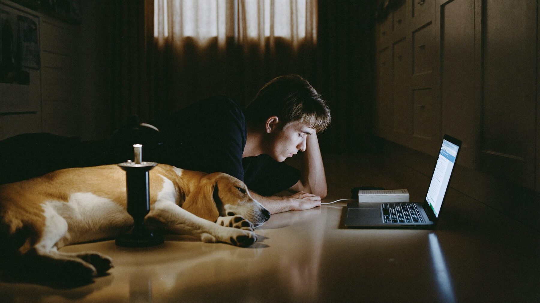 A man on a kitchen floor at night with a laptop and a sleepy beagle in his lap
